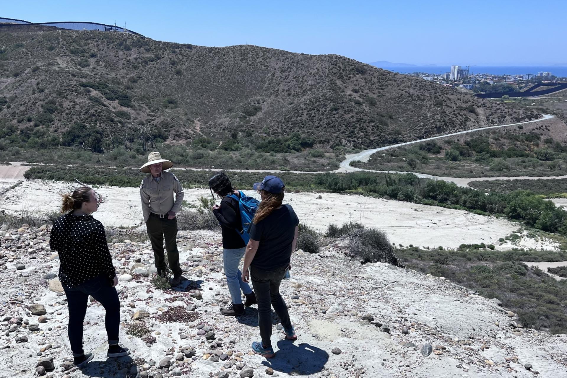 People standing relatively near the border wall between the United States and Mexico. There is a city in the background next to the Pacific Ocean.