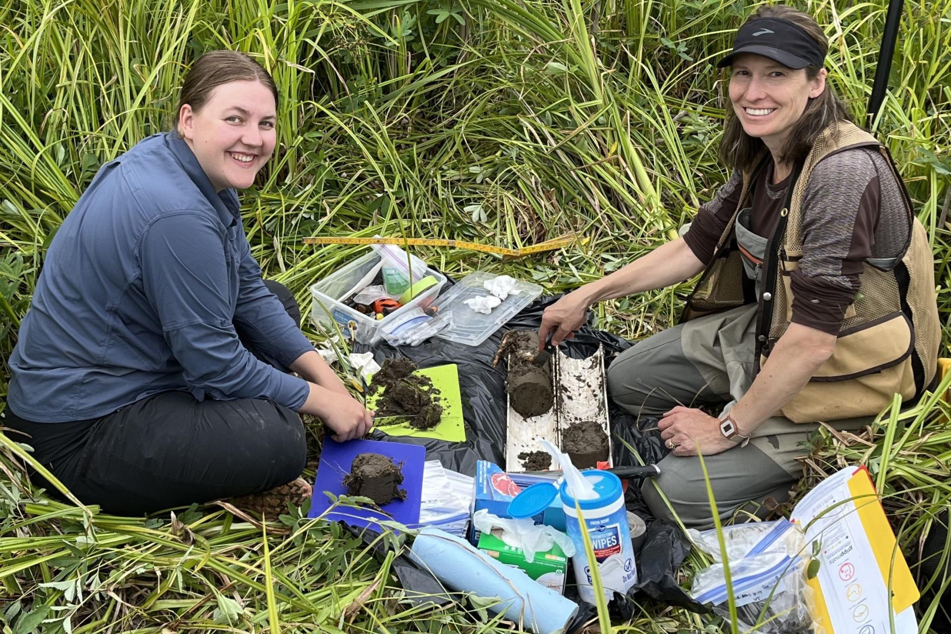Two individuals sitting on grass next to soil samples. One person is cutting into the soil with a basic kitchen knife.