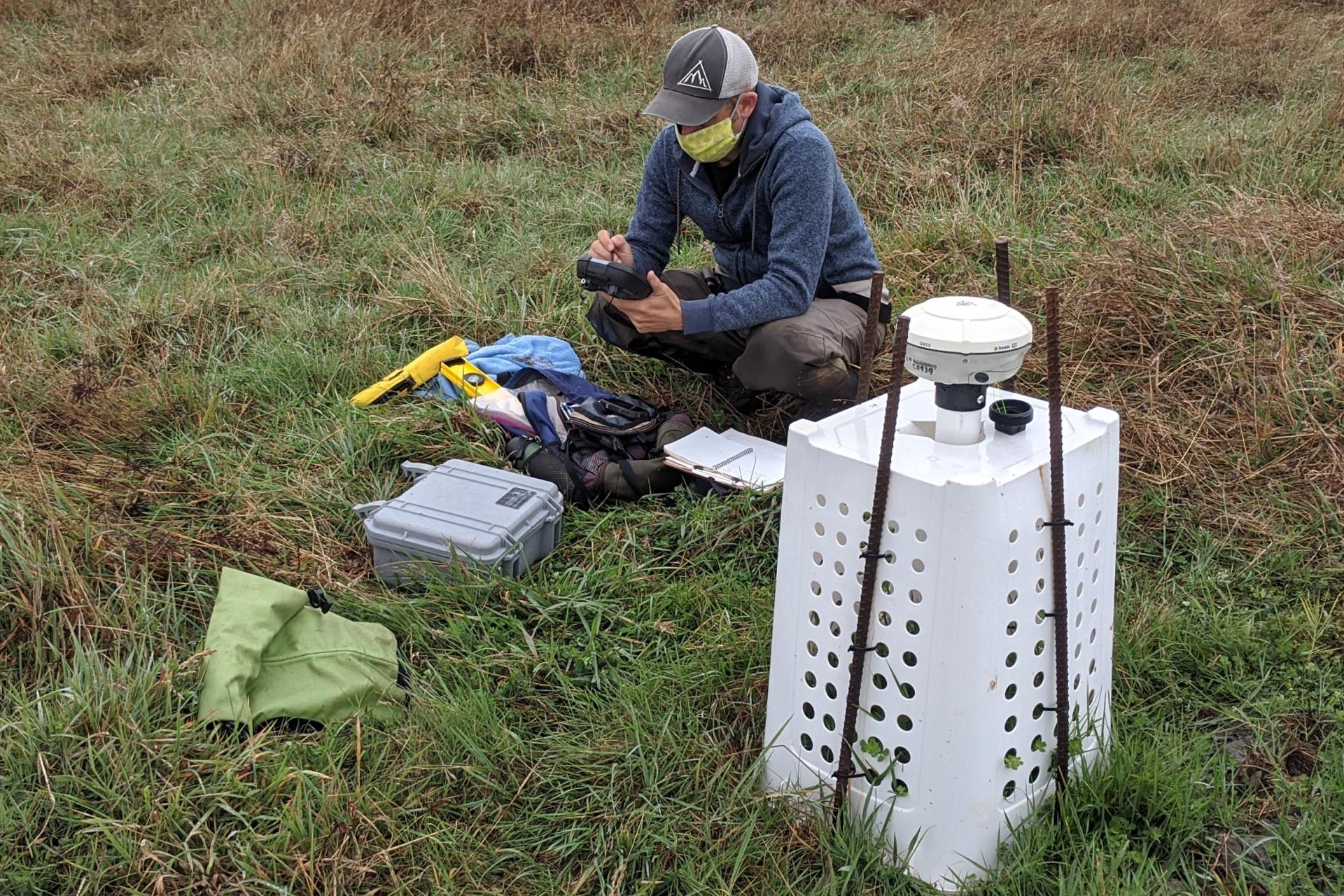 Person squatting and recording information on a hand-held device. There is a white instrument a few feet in front of the individual.