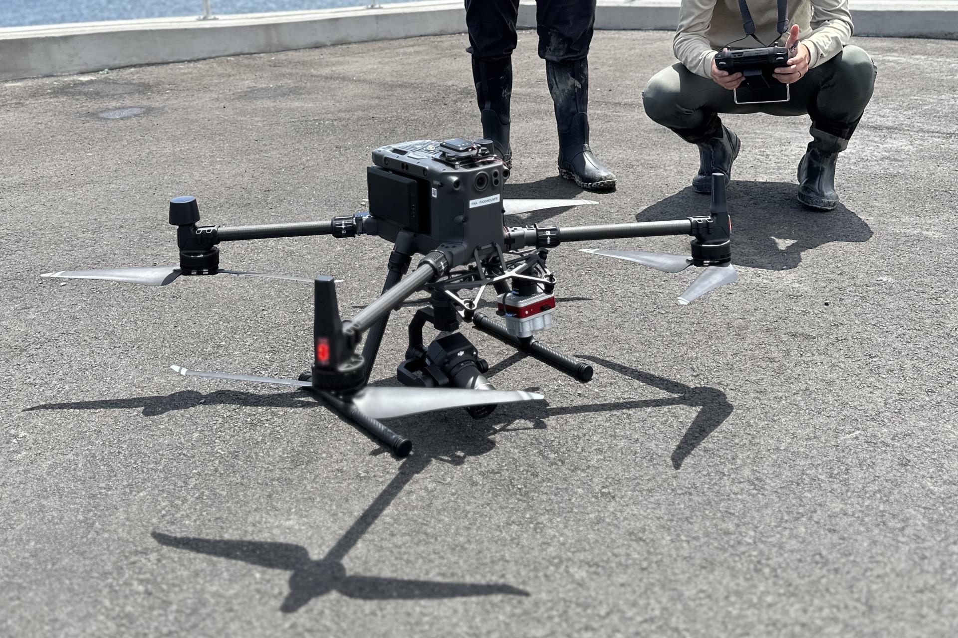 A person squatting holding a control for the drone. There is another person standing next to the drone operator.