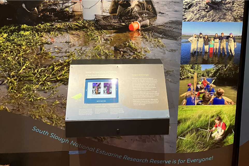 (3/4) An exhibit at the SSNERR visitor center displays the project findings. It features video simulations of estuarine temperature and salinity fluctuations generated by the project's hydrodynamic model. The model sits on a shelf that extends from the wall towards the viewer. The model shelf is surrounded by a collage images of the project, such as one being a group photo of people in waders and one being a group of students canoeing on a river with the viewer inside the canoe.