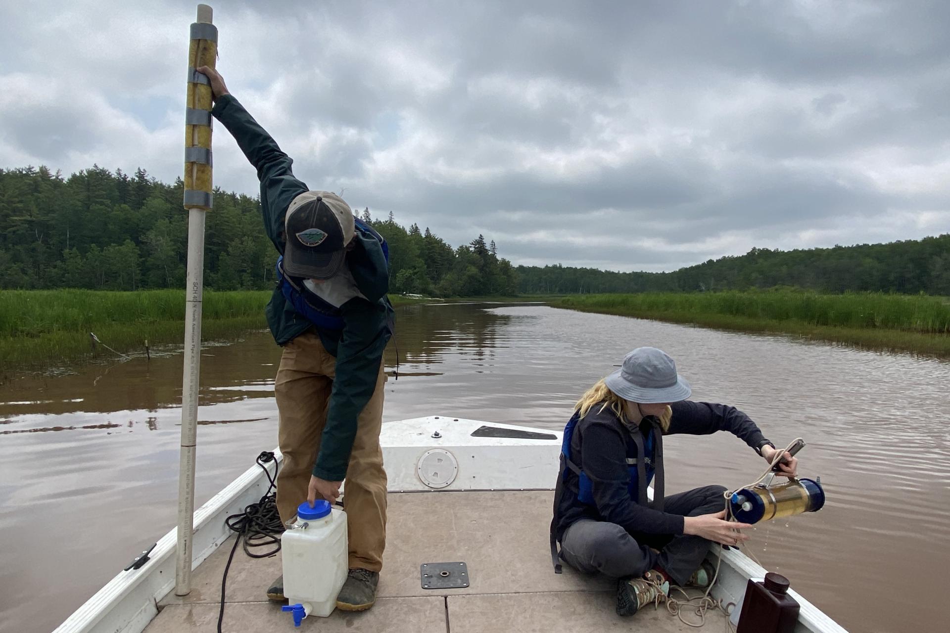Two people on a boat. One is standing with a large stick device and an empty canister. The other is sitting with a cylinder device filled with water.