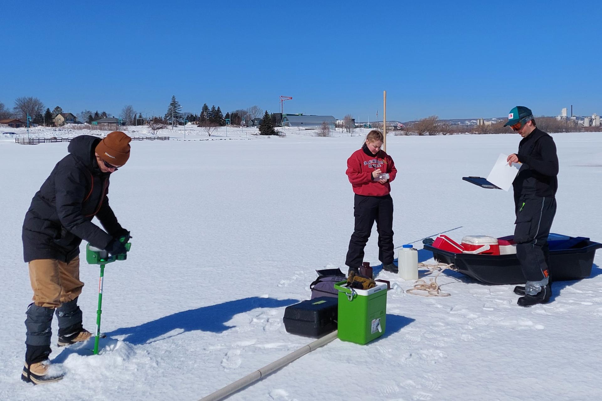 Three people on a frozen lake. The person on the left is making an hole in the ice using a green ice drill